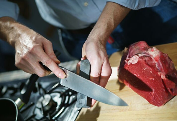 Hands hold a sharpener and knife angled on sharpener. A chunk of red meat and a gas range can be seen on the sides of the photo.