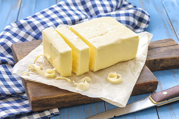 A block of butter features two cut pieces and some butter shavings on a white paper on top of a brown cutting board. A blue checkered towel and knife are off to the sides of the photo. The image image is on a blue background. 