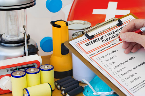 Clipcoard with emergency checklist, hand holding pen. Clipboard is surrounded by batteries, flash light, lantern, first aid kit