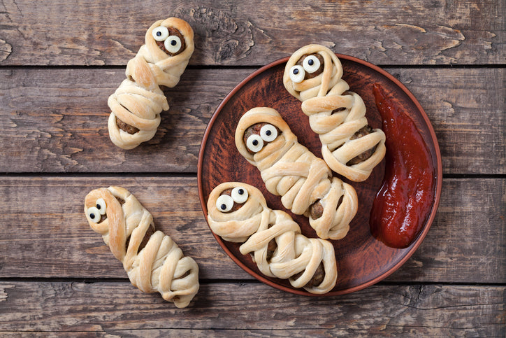 Food known as pigs in a blanket features hotdogs wrapped in shreds of dough, dressed to resemble mummies with candy eyes on a plate with ketchup, with a table background.