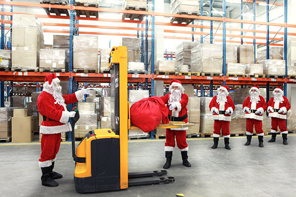 An image of five Santas in a warehouse. One Santa works a large machine while another hoists a red bag. The other three santas stand in line. 